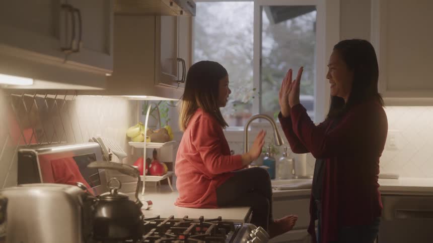 A child wearing a pink sweater sits on a kitchen counter giving a highfive to an adult standing nearby with various kitchen items and a window in the background