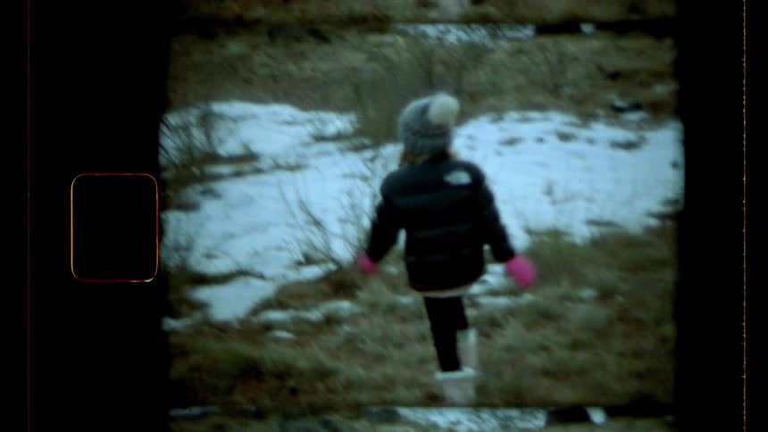 A little girl is running backwards in an environment that has a little snow on the ground and she is wearing warm winter clothes