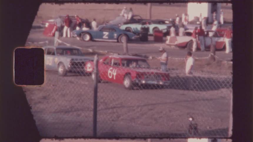 Stock cars are parked in the dirt as people walk around