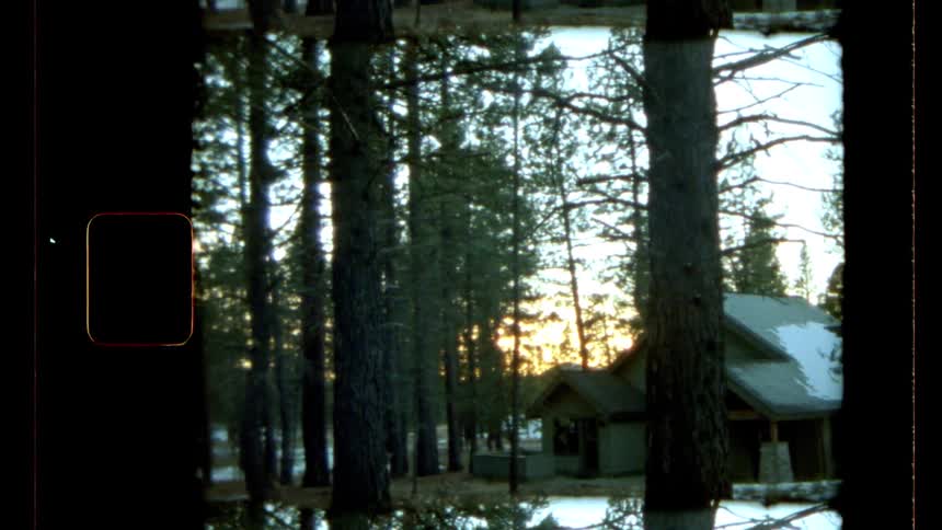 Snowy patches sit on the roof of a cabin and on the ground of the surrounding conifer forest as red tints the horizon in winter