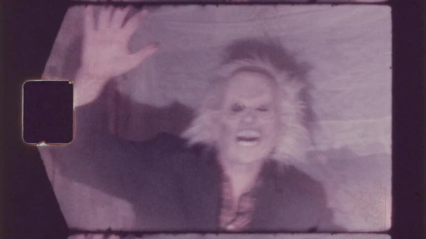 A person in a dark jacket stands with both hands raised wearing a halloween mask resembling a monster in front of a patterned curtain