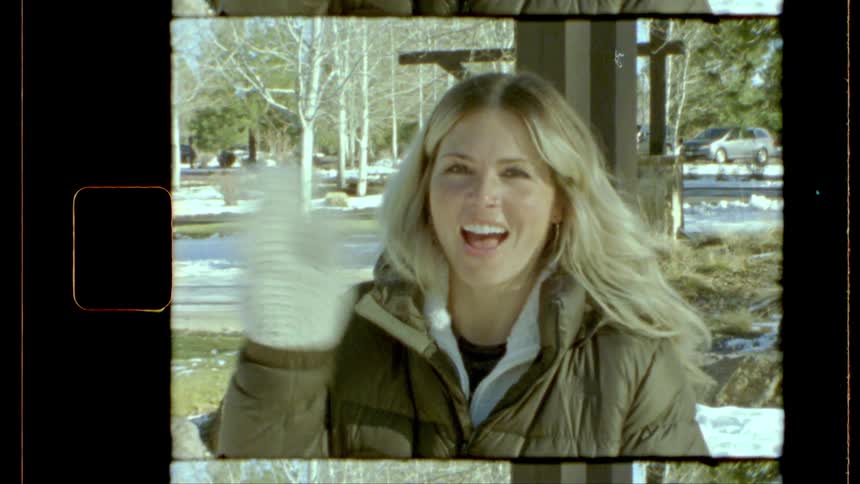 A blonde woman in a green jacket and white mittens waves for the camera in a snowy park with trees and a parked car in the background