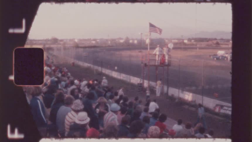 A vintagestyle shot shows a crowd gathered on tiered seating overlooking a racetrack with a race in progress