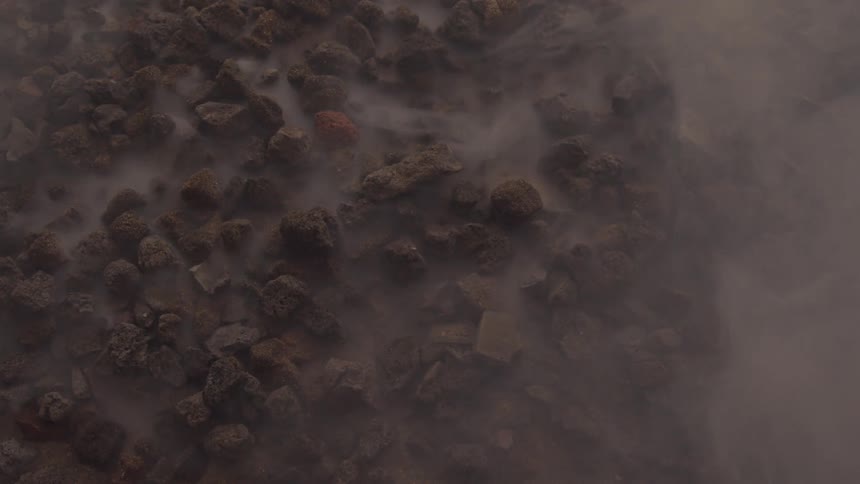 A rugged dark rocky surface with light mist or steam gently rising suggesting geothermal activity or heat