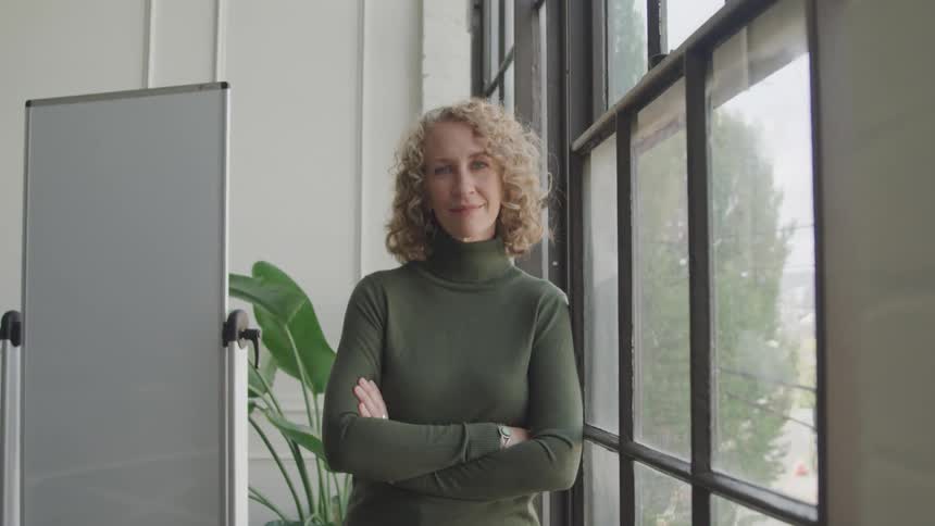 A woman with curly blonde hair dressed in a green sweater stands beside a window in an office
