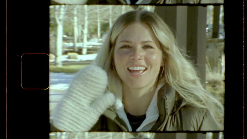 A smiling young blonde woman waves at the camera wearing white mittens and winter clothes outdoors