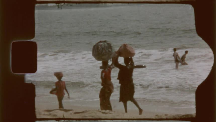 Three figures walk along a sandy beach towards the ocean with two carrying items on their heads captured on vintage film