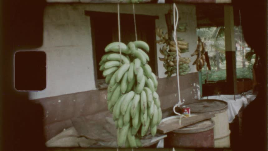 A bunch of green bananas hanging from a rope near a wall of a house with a person nearby