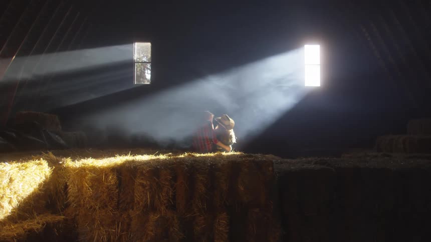 Couple sits in the hayloft and hugs while sunlight shines into the building through two windows