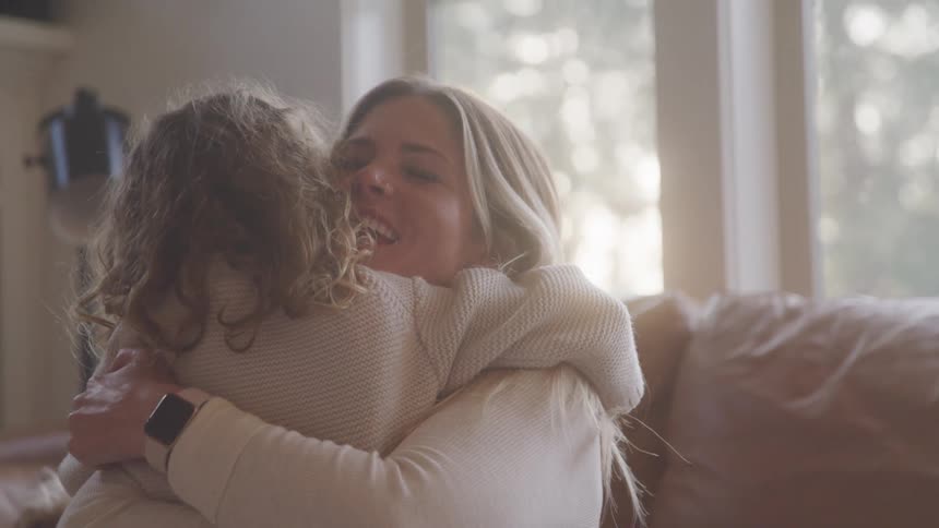 A mother hugs her daughter on a couch in a sunlit cozy living room near a window