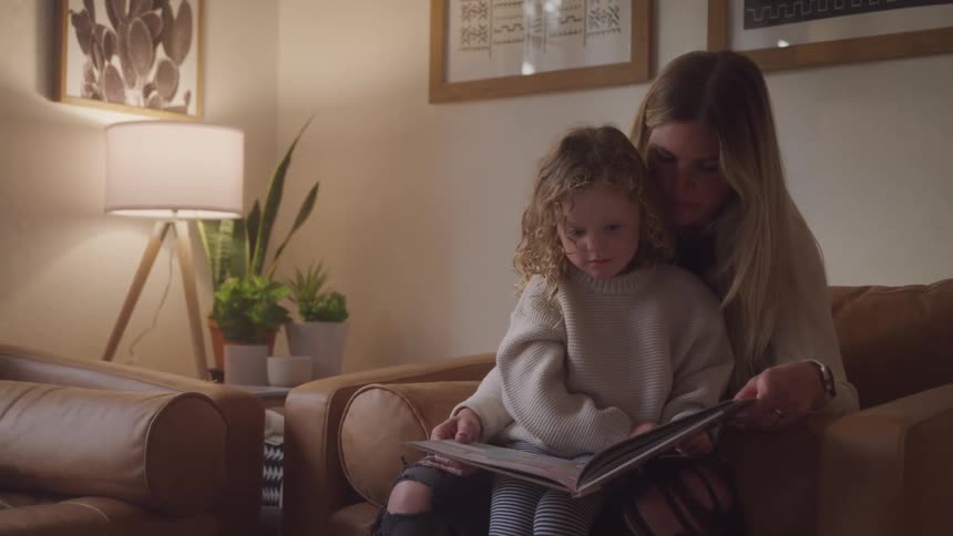 A mother and her daughter are sitting on an armchair while looking at pictures in a large book