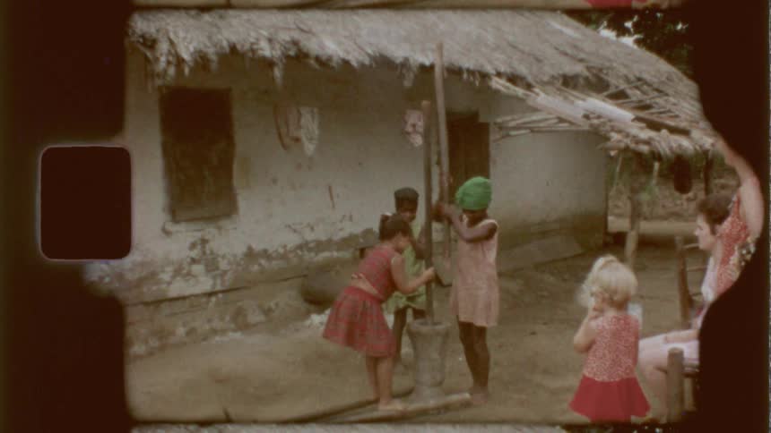 Children play and interact around a wooden pillar hammering on a tube while a woman and another child watch closely