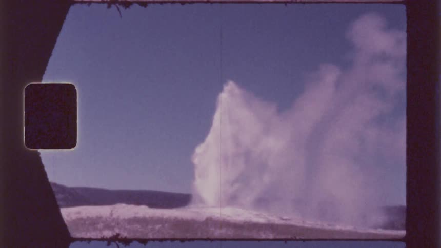A large plume of white vapor erupts against a clear blue sky partially obscuring a dark low lying landscape in the distance