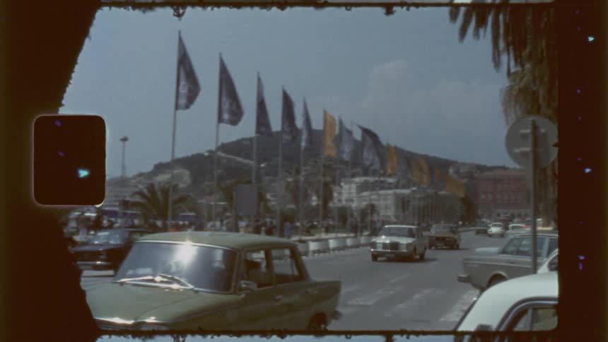 A street scene with classic cars driving towards the viewer framed by a mountain backdrop and a row of flags