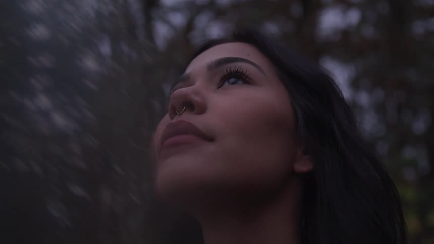 A young woman with dark hair and a nose ring gazes upward with a serene expression surrounded by softly blurred trees in the background at dusk