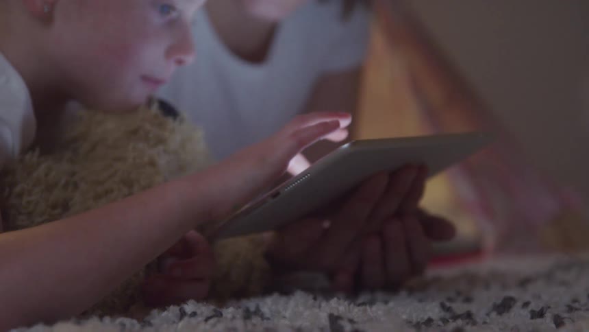 Little blonde girl lying on the bed interacting with a tablet