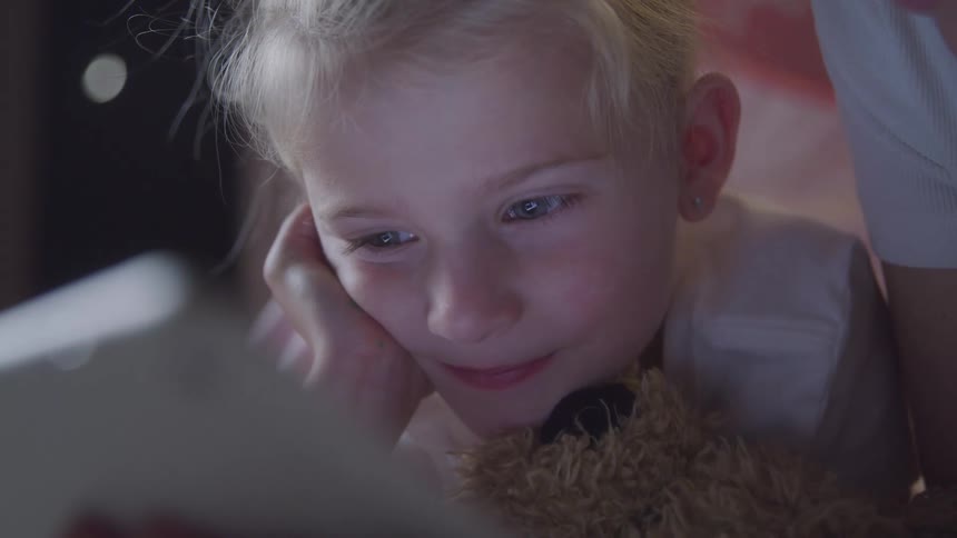 Little blonde girl with happy facial expression looking at a screen next to her mother