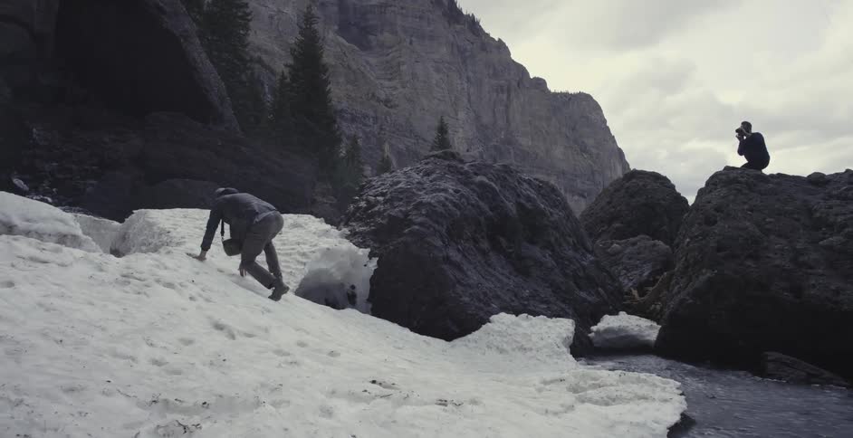 A man is crawling up some ice as another is on a rock photographing