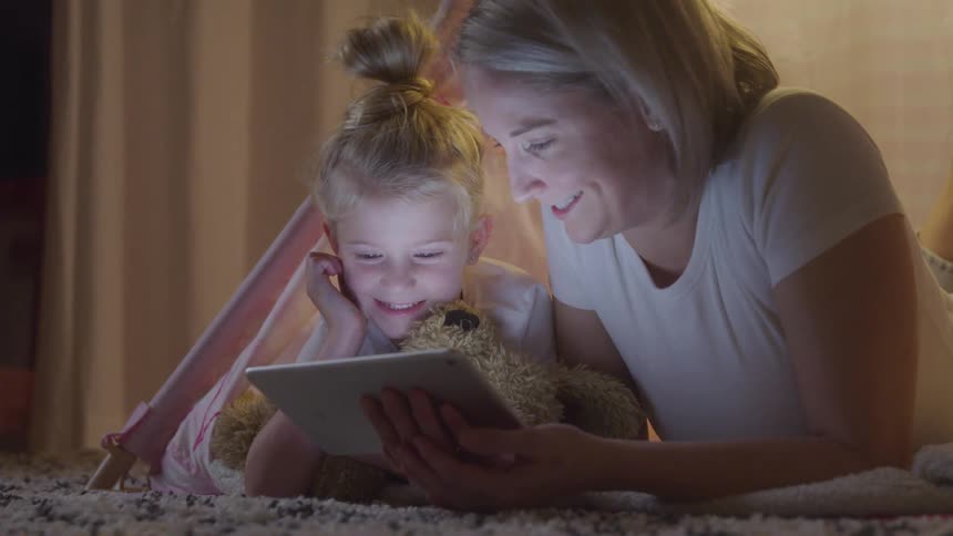 Mother and daughter lying face down on bed interacting with tablet with happy facial expression