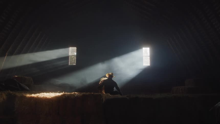 Two men in cowboy hats are sitting on hay in a dark building