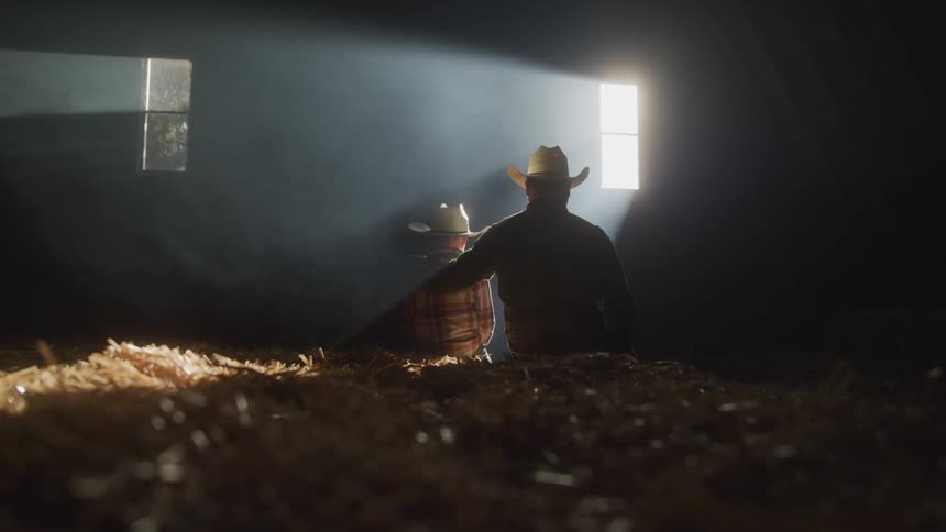 A father and child both in cowboy hats sit together in a sunlit barn silhouetted against the light