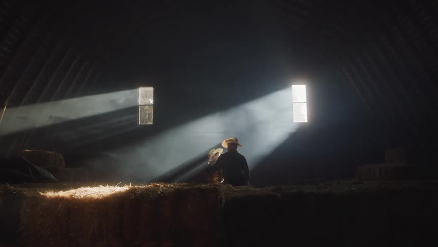 Father and son sitting in a barn lit by rays of light coming through the windows