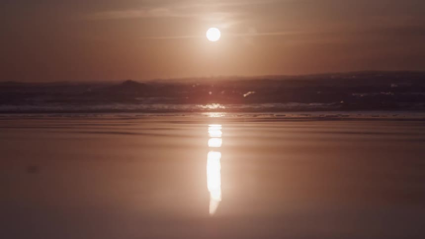 The suns reflection creates a shimmering vertical line on the wet sand of a beach at either sunrise or sunset