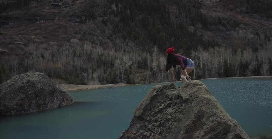 A woman stands on top of a large rock near a body of water