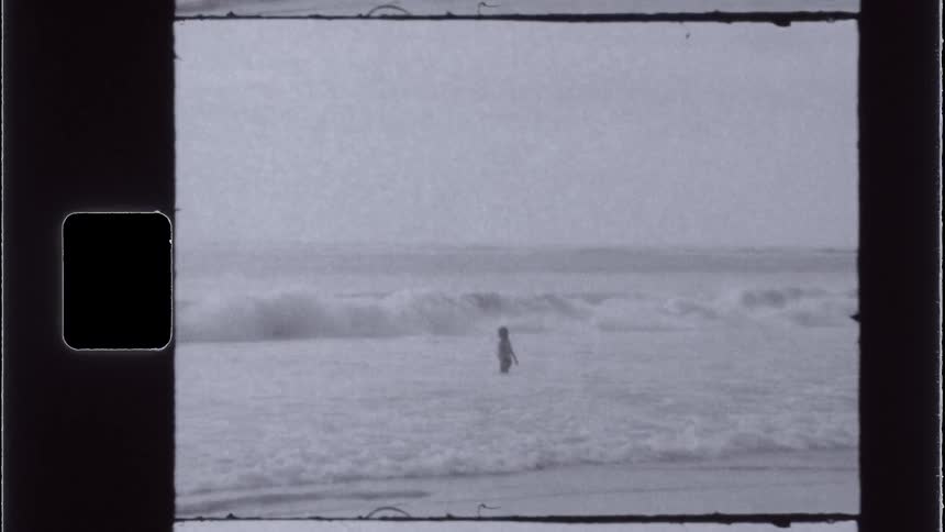 A vintagestyle shot shows a lone figure standing in the ocean surf with waves crashing around them under an overcast sky