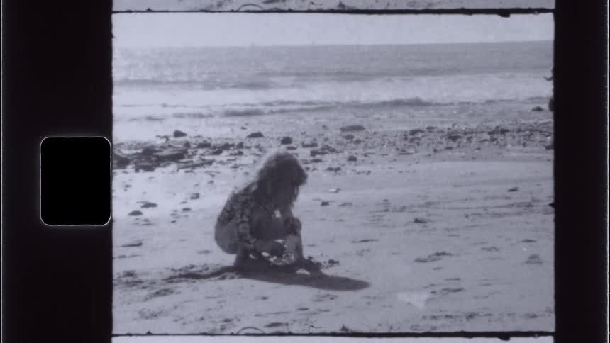 A vintagestyle shot shows a person crouching on a sandy beach playing or examining something in the sand with the ocean visible in the background