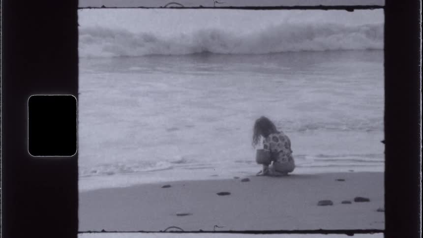 A vintagestyle shot shows a girl kneeling on a sandy beach at the waters edge with waves visible in the background