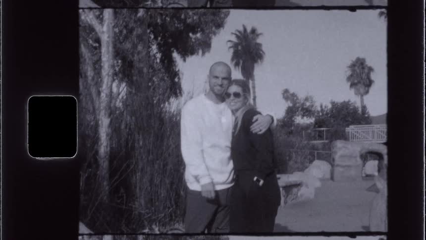 A vintagestyle shot shows two people standing closely together with one persons arm around the other in an outdoor setting with trees and palm trees in the background