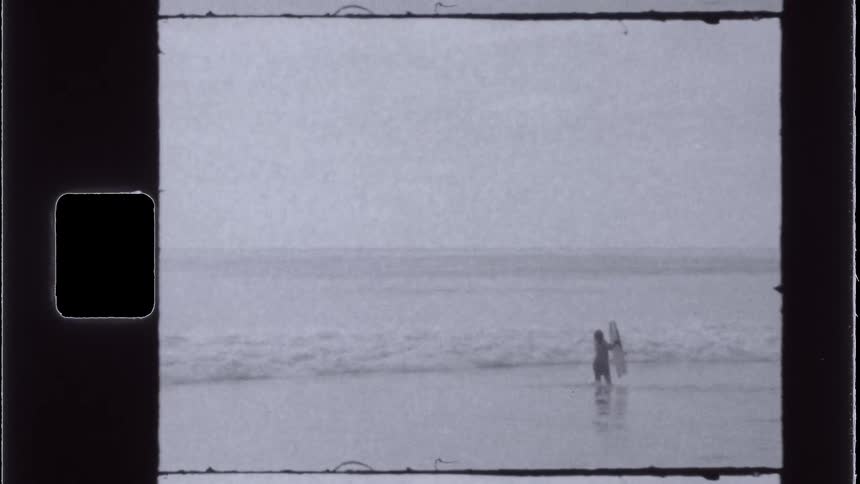 A vintagestyle shot shows a lone figure carrying a surfboard standing in the ocean surf with waves crashing around them under an overcast sky
