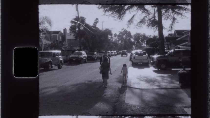 An adult and two children walking on the edge of an asphalt street with several parked cars and everything is in black and white tones