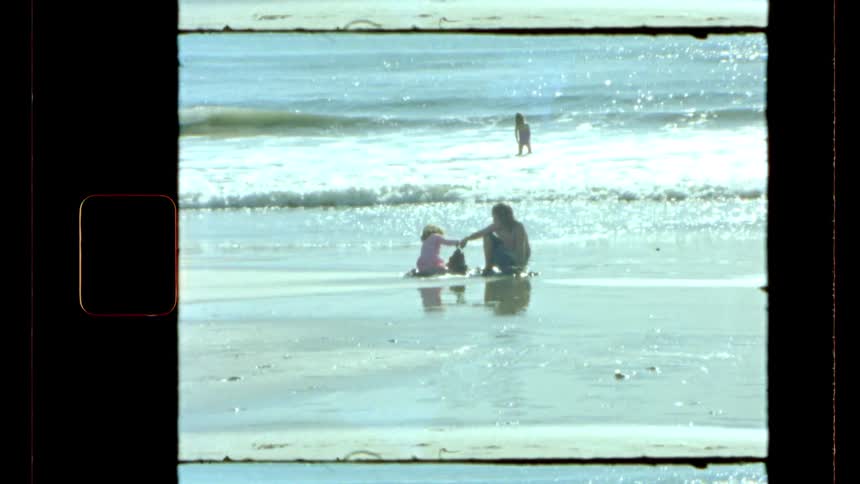 A vintagestyle shot shows two people sitting on a wet sandy beach playing at the waters edge
