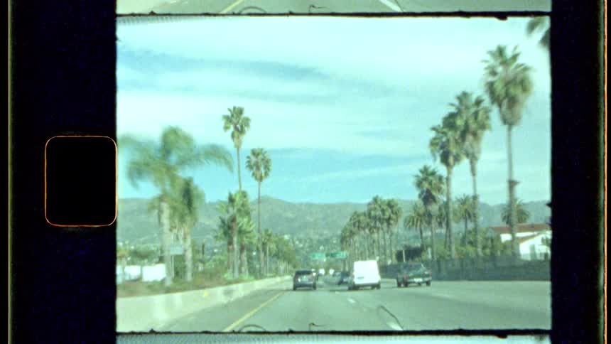 A vintagestyle shot shows a highway scene with several vehicles driving past palm trees and distant hills under a light sky