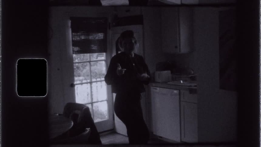A woman in a kitchen stopping to strike a pose with her arms and everything is in black and white tones