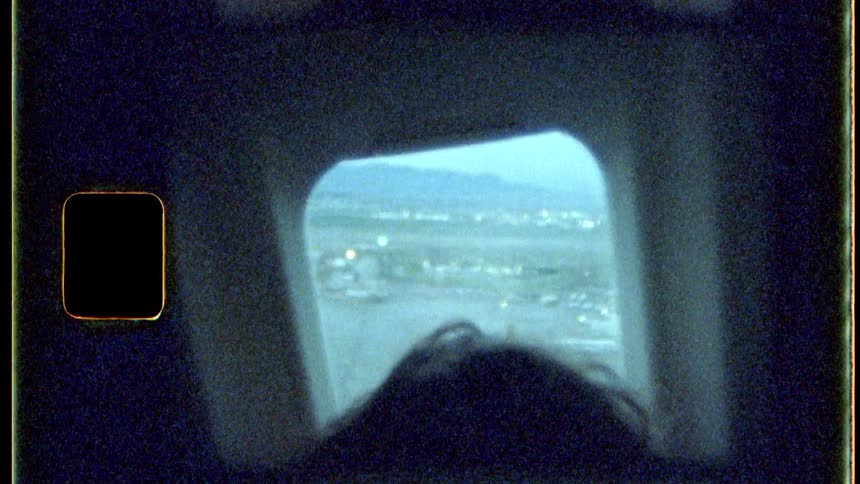 A vintagestyle shot shows an airplane window framing a distant city skyline and mountains with the back of a persons head visible in the foreground