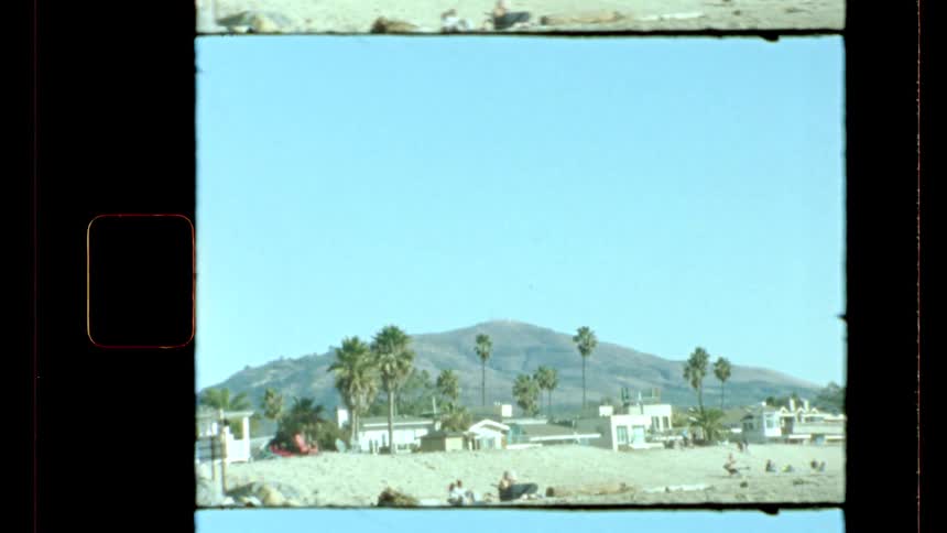 A vintagestyle shot shows a coastal scene with buildings and palm trees in the foreground a hill in the midground and a clear sky