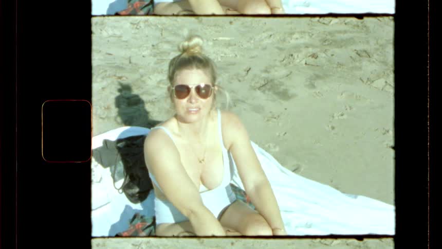 A vintagestyle shot shows a woman wearing sunglasses and a swimsuit sitting on a beach towel smiling at the camera