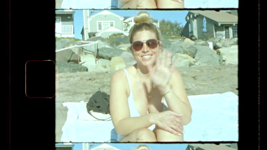 A vintagestyle shot shows a woman wearing sunglasses and a swimsuit sitting on a beach towel smiling at the camera