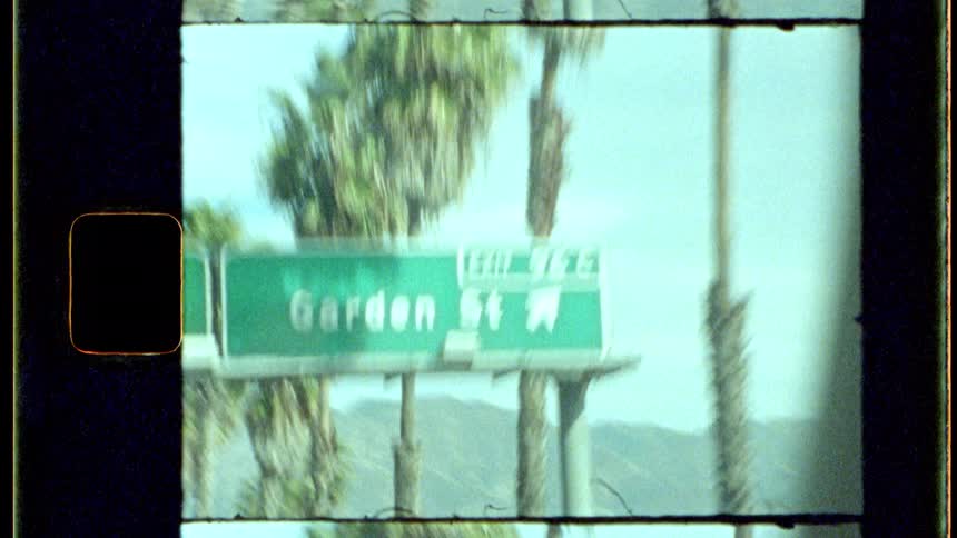 Through the window of a moving vehicle bright green highway signs are prominently displayed standing in contrast to the towering palm trees lining the roadside and a clear bright sky above all presented with the aesthetic qualities of analog film