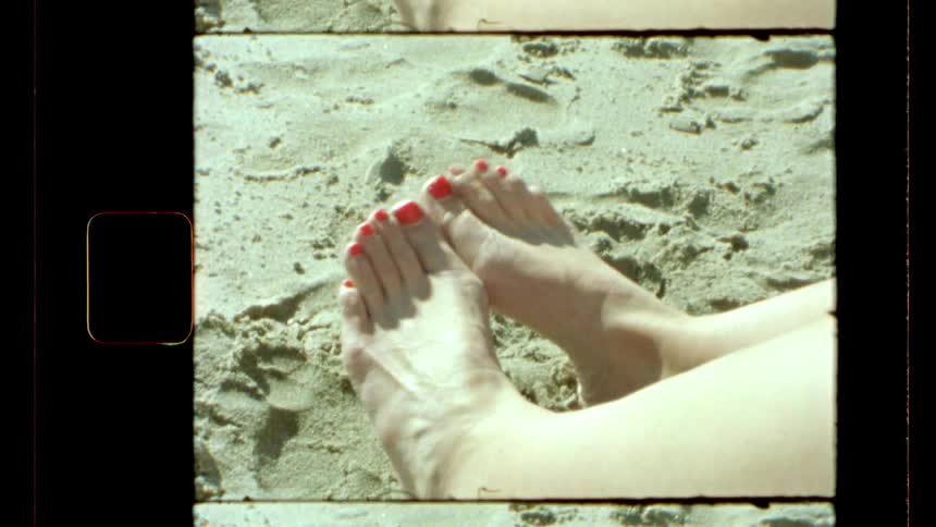 A vintagestyle shot shows two bare feet with red painted toenails resting on sandy ground