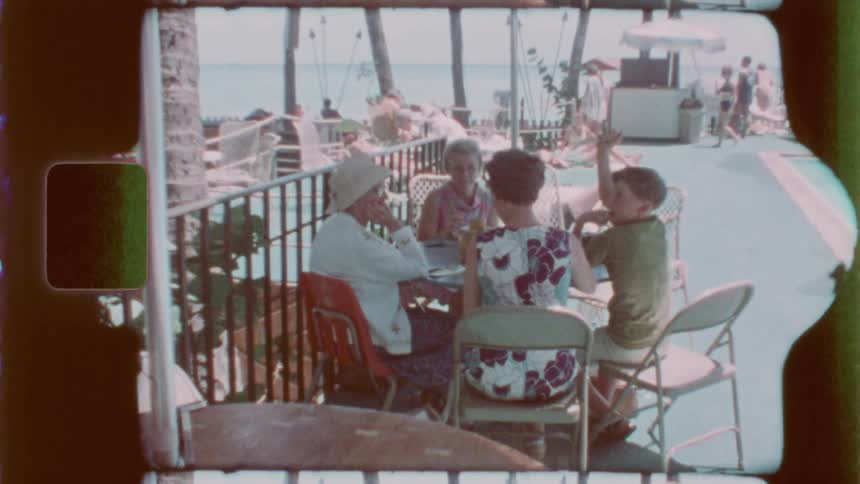 A vintagestyle shot shows family gathered around a table at a resort or vacation destination near the beach