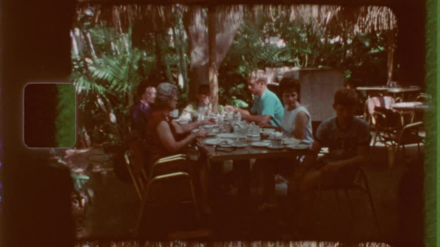 A vintagestyle shot shows several people gathered around a table outdoors dining amidst lush foliage