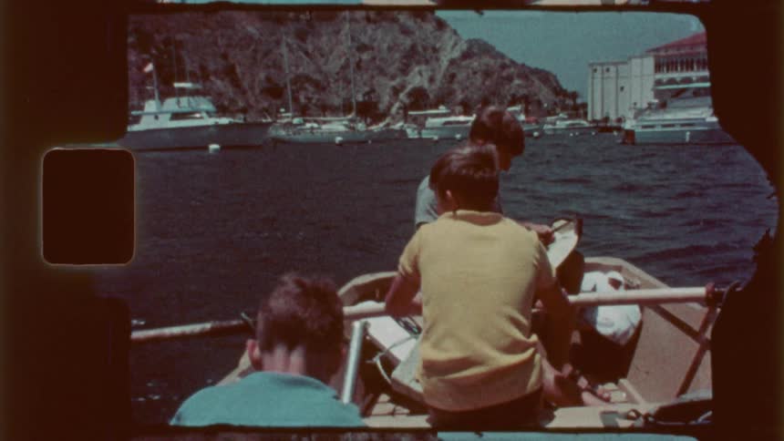 Three boys are in a small boat while one rows with the oars toward other boats and a marina