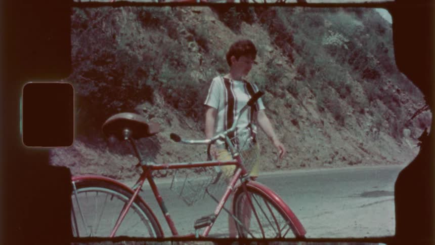 A teenage boy walking on a rural asphalt street next to a bicycle and a mountain behind