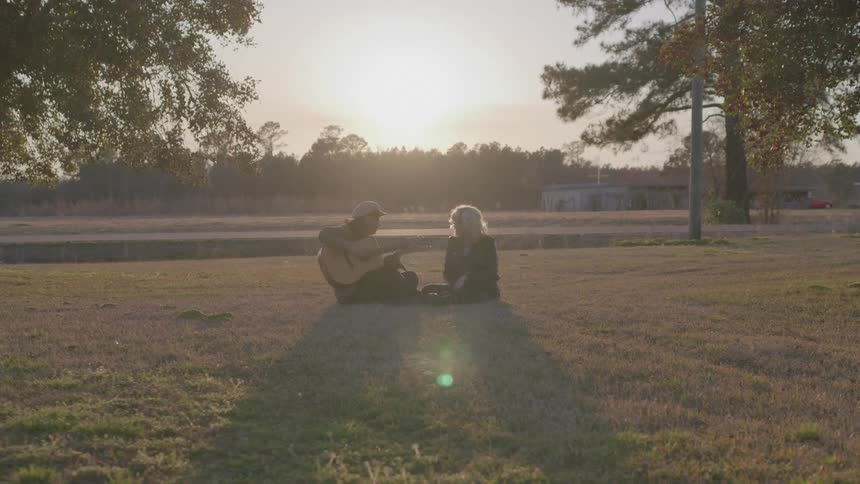 Two people sit on grass in a field at sunset with one playing a guitar and the other listening surrounded by trees on a park near a road