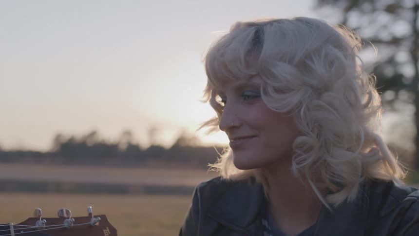 A woman outside smiles and looks at the neck of a guitar