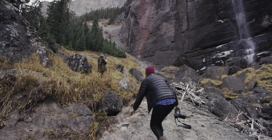 A woman carries a camera and walks up a hill to another person standing in the mountains